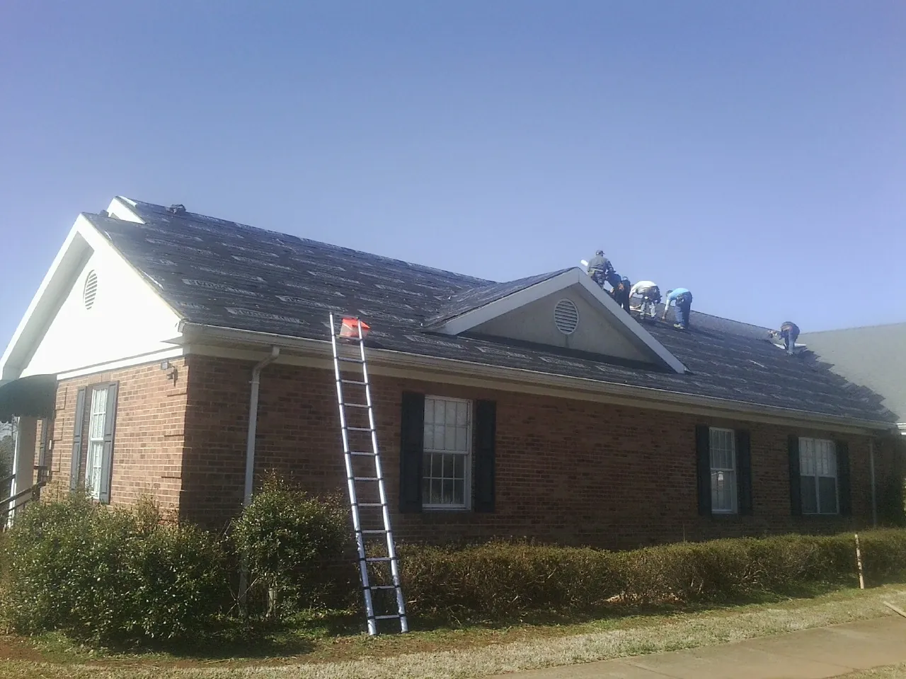 Crew tearing shingles off a brick ranch roof with a ladder set — Emergency Roof Repair work by our crew in Wood River Junction