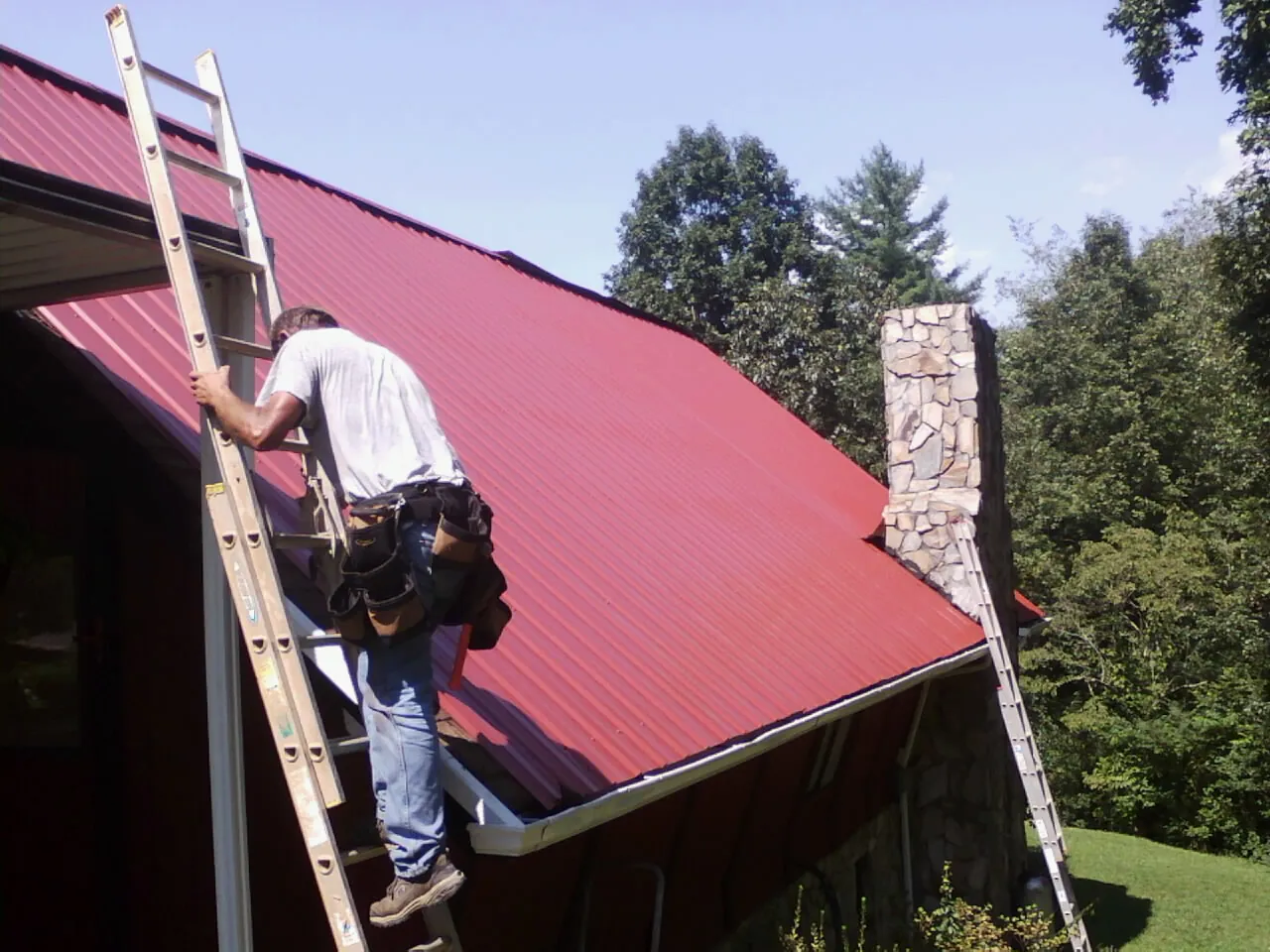 Roofer on a ladder beside a red metal roof and stone chimney — Soffit & Fascia Repair by Roofing Brothers in Wood River Junction